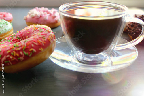 Glass Cup of hot coffee on a black table surrounded by colorful donuts close-up