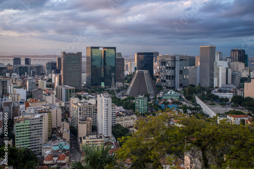 High vantage view of the city centre of Rio de Janeiro with skyscrapers and the cone shaped cathedral in the middle at sunset with dramatic clouds overhead © Maarten Zeehandelaar