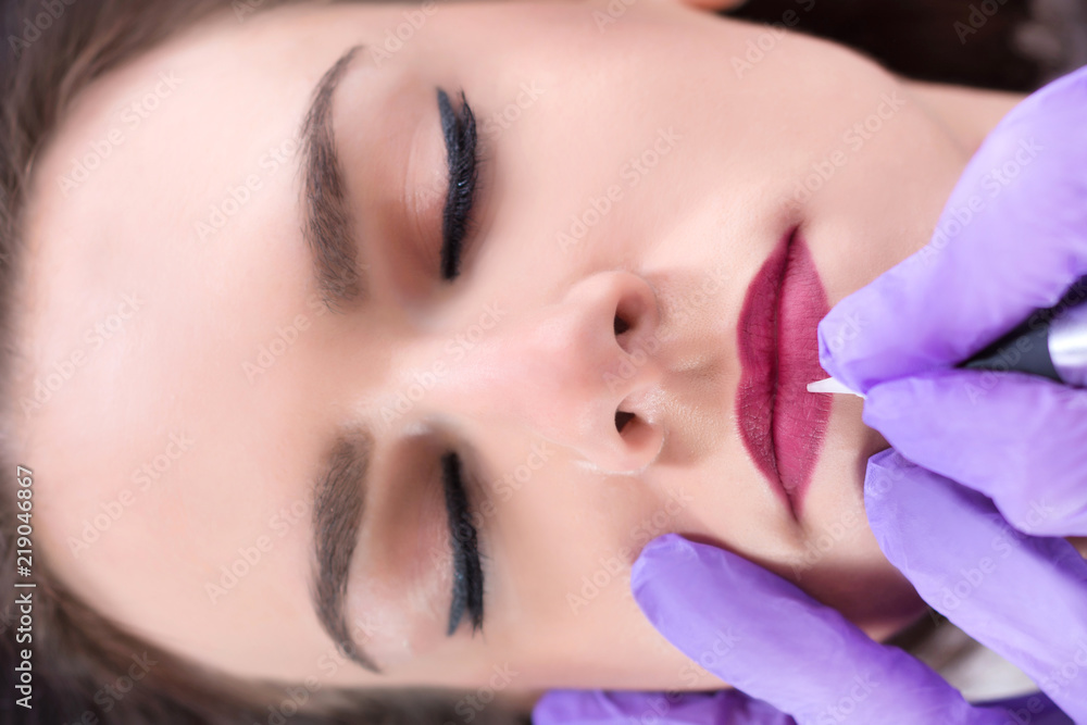 Applying red permanent make up tattoo on young girl lips in beauty salon. Modern and new make up concept. Close up, selective focus