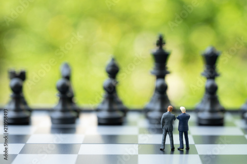 Partnership and teamwork in business strategy concept, two miniature people businessmen work as team, standing on chessboard looking at black chess in far background, finding solution for success