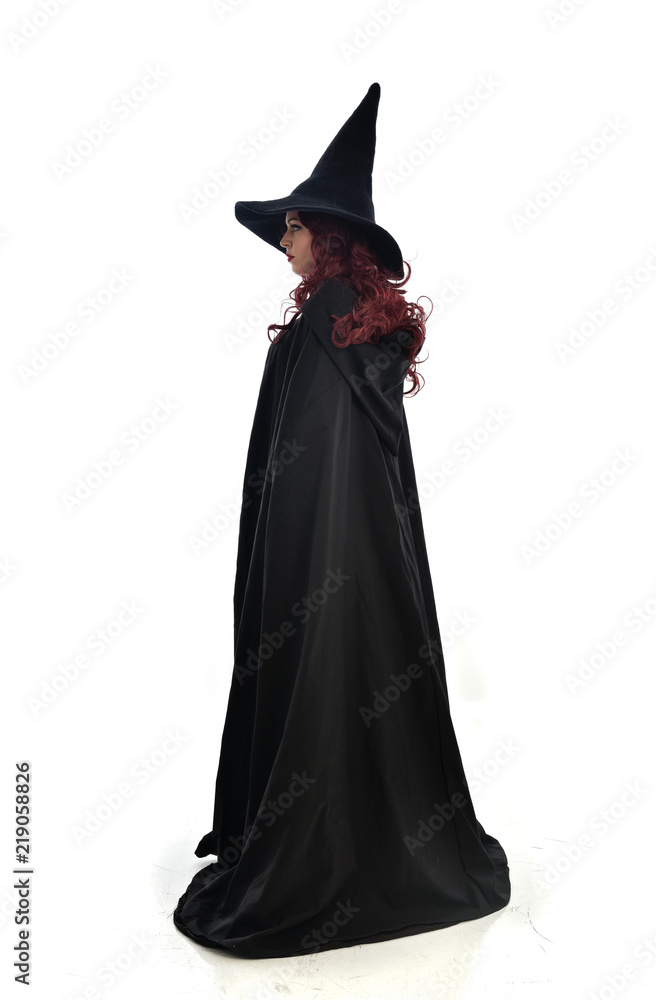 full length portrait of red haired girl wearing long black cloak, pointy hat and witch costume. standing pose, isolated on white studio background. full length portrait of red haired girl wearing long black cloak, pointy hat and witch costume. standing pose, isolated on white studio background.