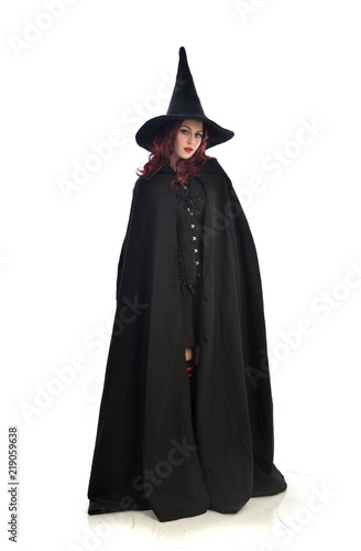 full length portrait of red haired girl wearing long black cloak, pointy hat and witch costume. standing pose, isolated on white studio background.