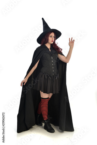 full length portrait of red haired girl wearing long black cloak, pointy hat and witch costume. standing pose, isolated on white studio background.