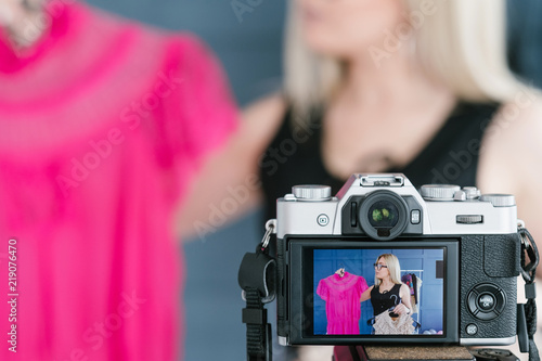 image of clothing designer advertising her new collection on the camera screen. concept of live video streaming trend in business. woman holding two tops in hands and vlogging.