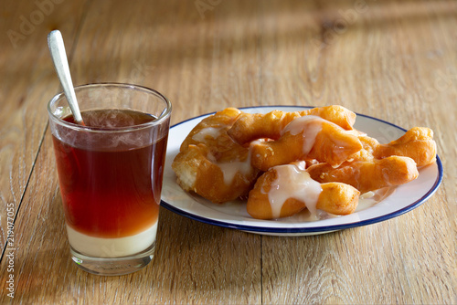 Hot tea with condensed milk in glass and deep-fried dough stick (Chinese Language Pa tong ko ) in white dish on wooden floor thai tea