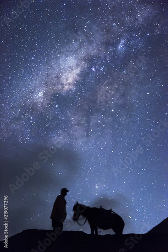 Horsemen standing under million stars in sky with his horse near smoking hot volcano Mount bromo at night, Indonesia