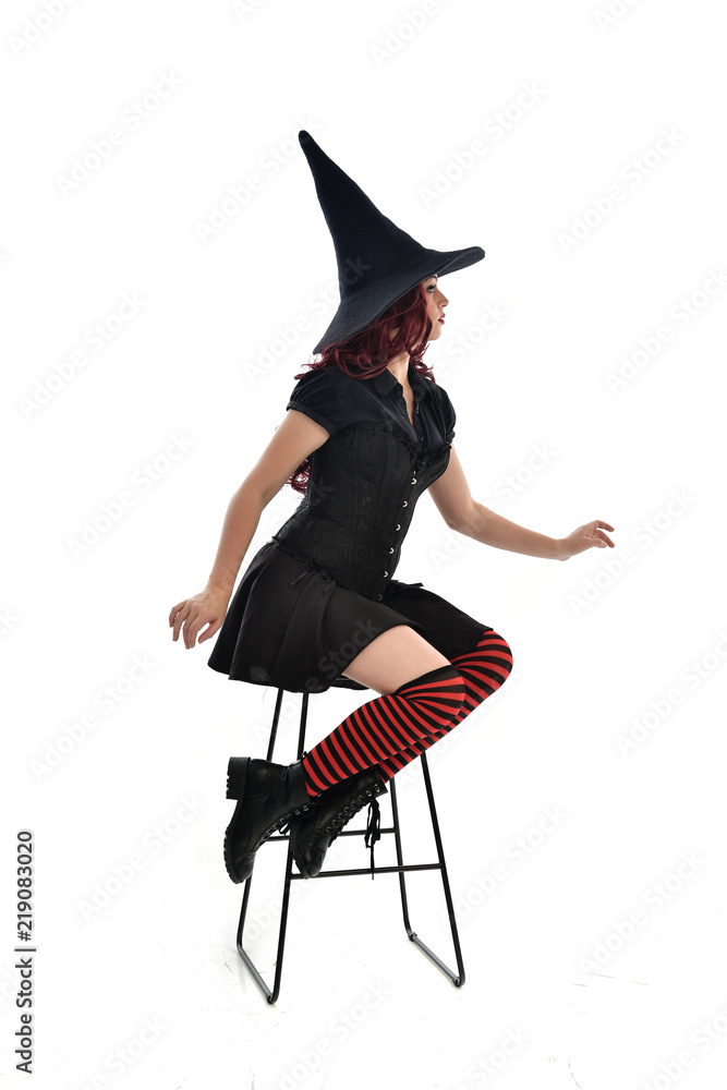 Obraz premium full length portrait of red haired girl wearing long black cloak, pointy hat and witch costume. seated pose on chair, isolated on white studio background.