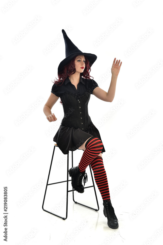 full length portrait of red haired girl wearing long black cloak, pointy hat and witch costume. seated pose on chair, isolated on white studio background. full length portrait of red haired girl wearing long black cloak, pointy hat and witch costume. seated pose on chair, isolated on white studio background.