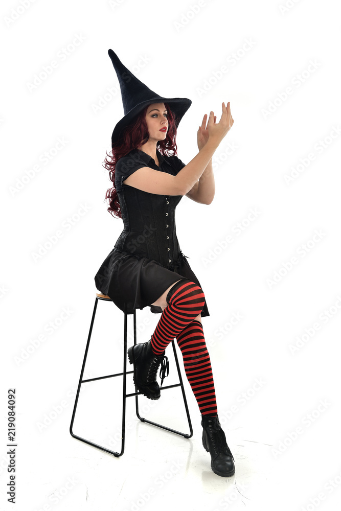 full length portrait of red haired girl wearing long black cloak, pointy hat and witch costume. seated pose on chair, isolated on white studio background. full length portrait of red haired girl wearing long black cloak, pointy hat and witch costume. seated pose on chair, isolated on white studio background.