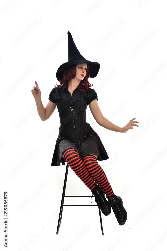 Obraz premium full length portrait of red haired girl wearing long black cloak, pointy hat and witch costume. seated pose on chair, isolated on white studio background.