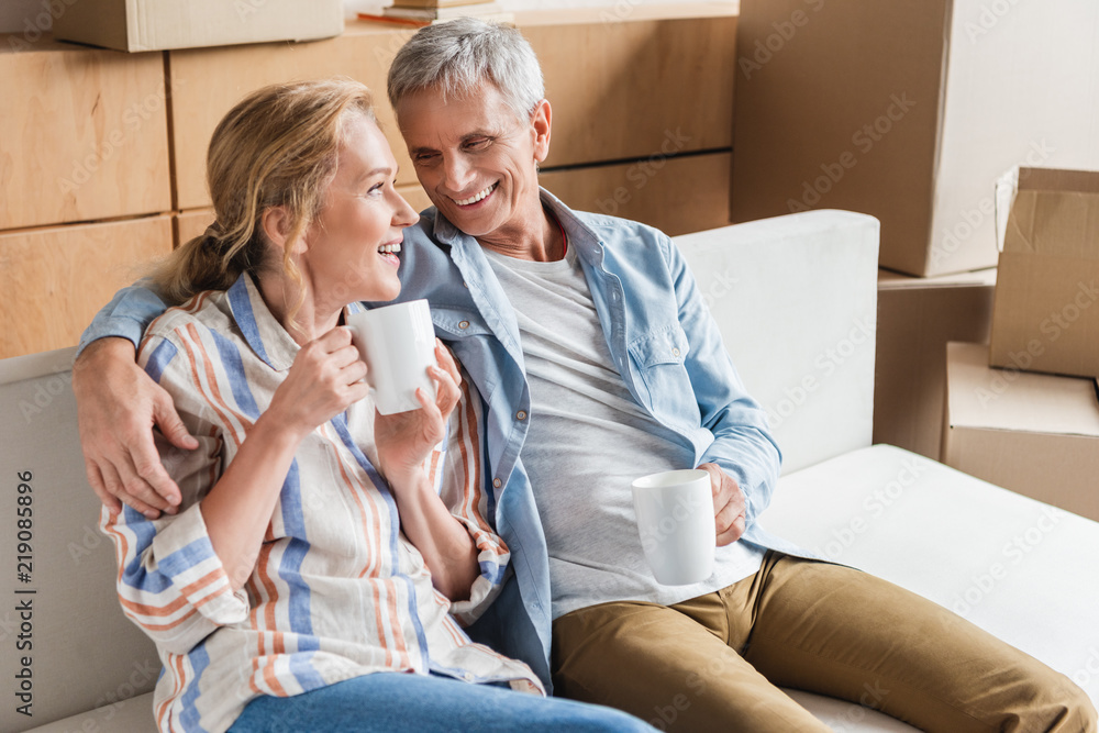 happy senior couple drinking tea and smiling each other while sitting on couch during relocation happy senior couple drinking tea and smiling each other while sitting on couch during relocation