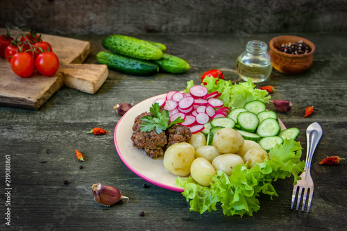 Home made meatballs are served with young potatoes, salad, sliced radishes and cucumbers. In a plate on a wooden table.