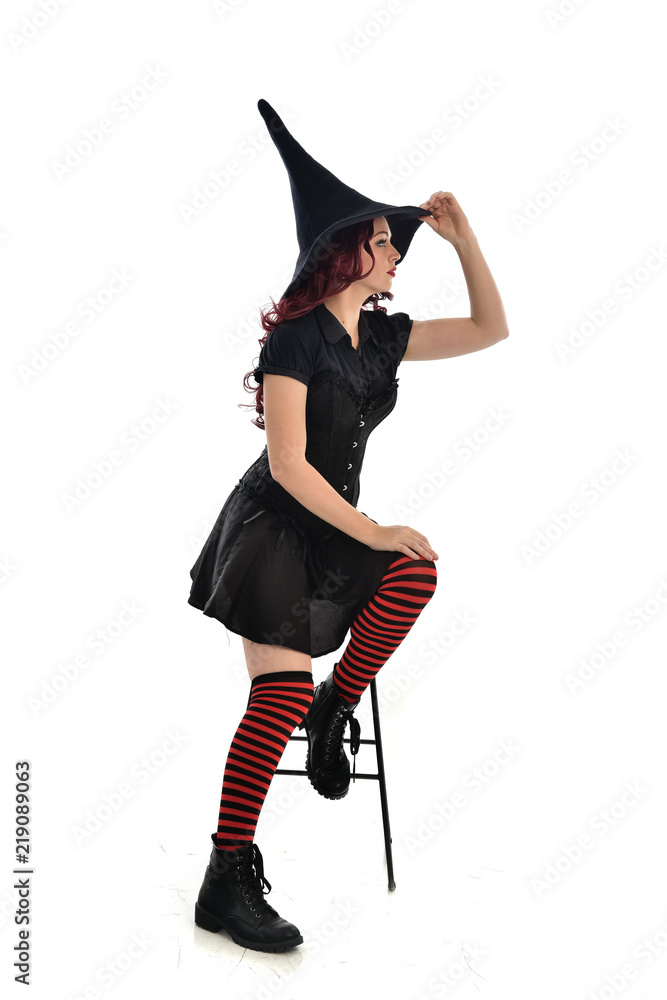 full length portrait of red haired girl wearing long black cloak, pointy hat and witch costume. seated pose on chair, isolated on white studio background. full length portrait of red haired girl wearing long black cloak, pointy hat and witch costume. seated pose on chair, isolated on white studio background.