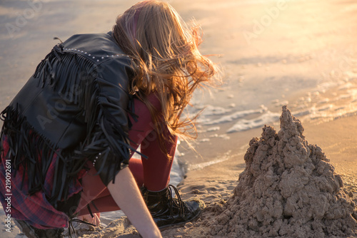 Young woman building and making sand castle next to sea. Calm, relaxing and romantic evening during sunset at the beach side. Remembering the childhood. Loneliness concept. 