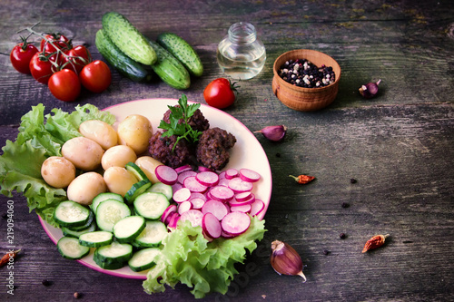 Home made meatballs are served with young potatoes, salad, sliced radishes and cucumbers. In a plate on a wooden table.