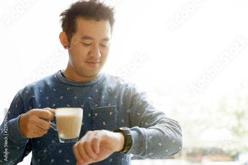 Cheerful asian man with coffee cup in hands watching on watches while standing in his home office in morning. Coffee break concept, Copy space. free from work and duty.