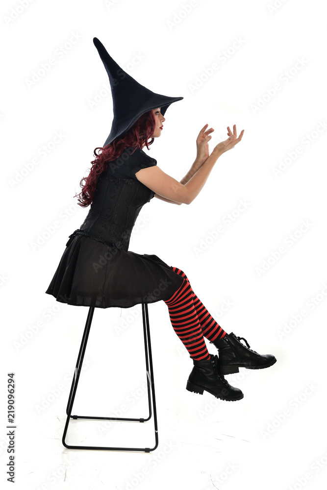 Obraz premium full length portrait of red haired girl wearing long black cloak, pointy hat and witch costume. seated pose on chair, isolated on white studio background.