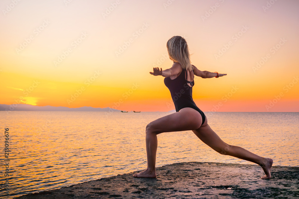 Beautiful girl in black doing yoga on the pier by the sea, standing asana virabhadrasana at sunrise. Healthy lifestyle.