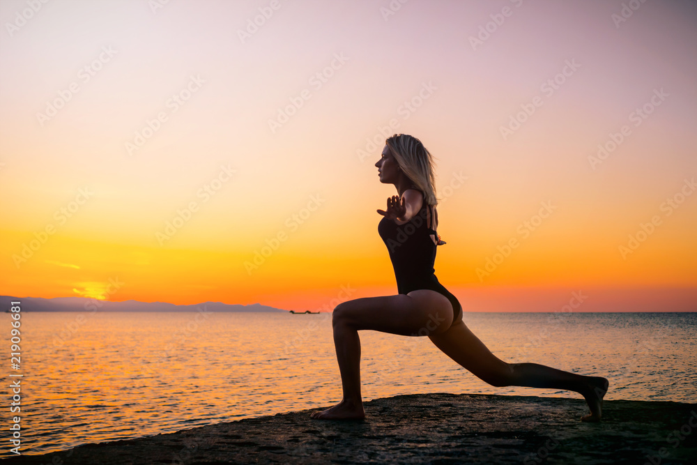 Beautiful girl in black doing yoga on the pier by the sea, standing asana virabhadrasana at sunrise. Healthy lifestyle.
