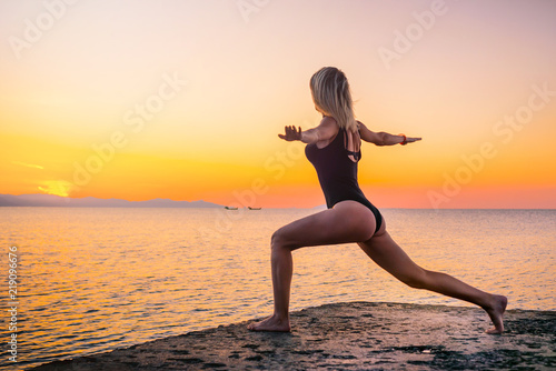 Fototapeta Naklejka Na Ścianę i Meble -  Beautiful girl in black doing yoga on the pier by the sea, standing asana virabhadrasana at sunrise. Healthy lifestyle.