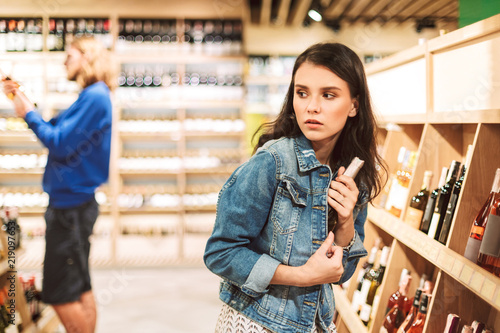 Young frightened woman in denim jacket trying steal bottle of wine in modern supermarket photo