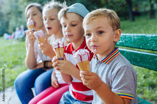 Group of four happy children eating ice cream together outdoor. Photo of happy blond girls with two handsome boys sitting on the bench and smiling at camera.
