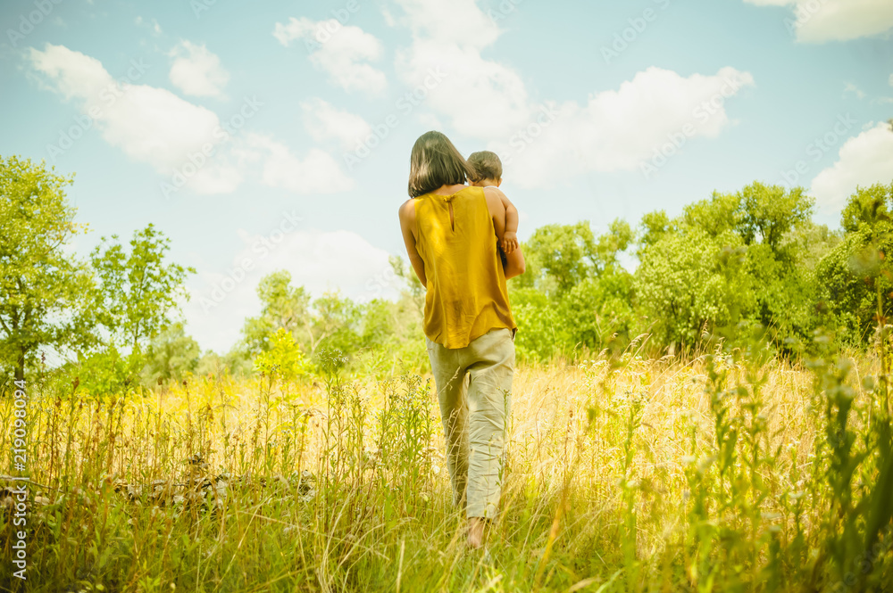 back view of mother holding son and walking in field at sunny day