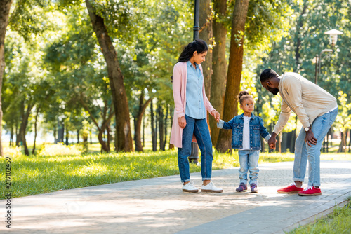 african american parents and daughter holding hands in park, father looking at daughter