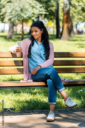 cheerful beautiful african american woman looking at smartphone on wooden bench in park