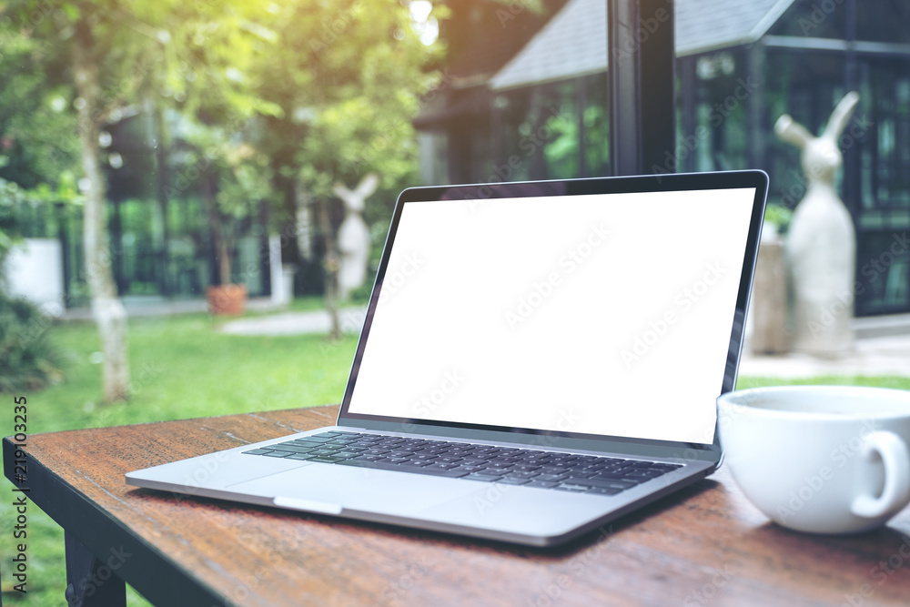 Mockup image of laptop with blank white desktop screen and coffee cup on wooden table with nature background Mockup image of laptop with blank white desktop screen and coffee cup on wooden table with nature background