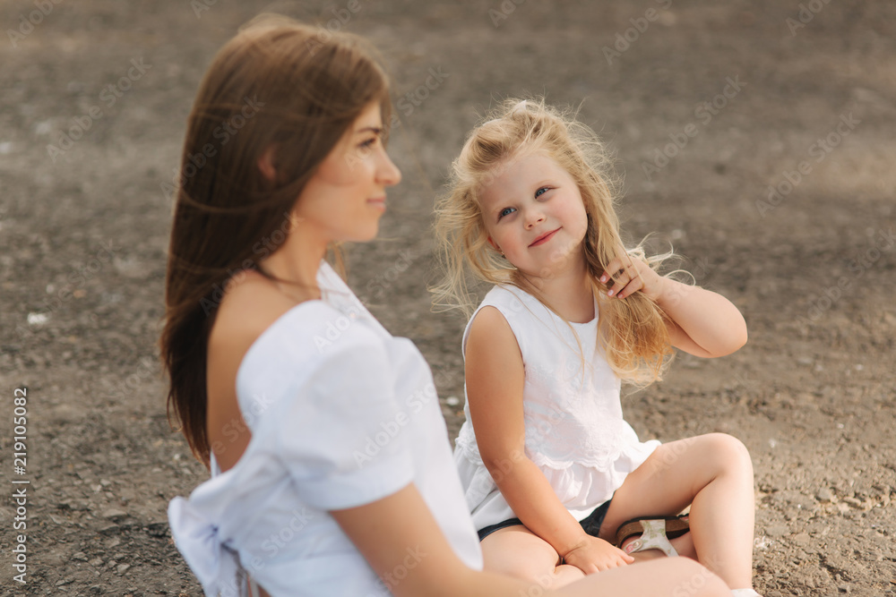Attractive Mom and blonde hair daughter sits on road near big alley. They smile and look to natune Attractive Mom and blonde hair daughter sits on road near big alley. They smile and look to natune