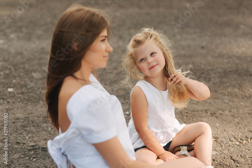 Attractive Mom and blonde hair daughter sits on road near big alley. They smile and look to natune