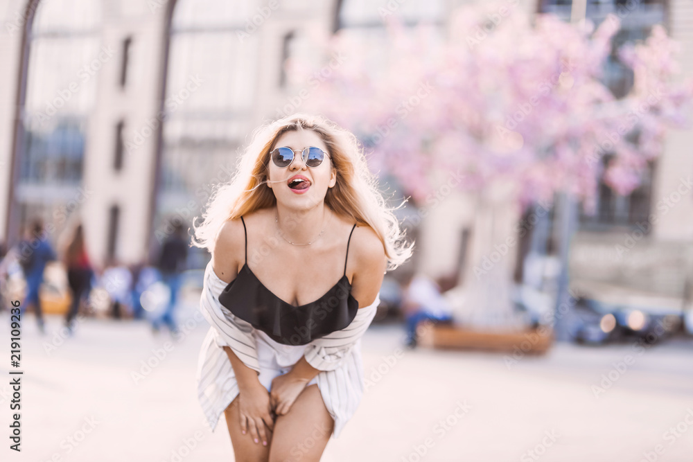 Portrait Beautiful young blonde girl in sunglasses with puffy lips and sexy feminine body posing on the street with pink flowers on the background on sunny day Portrait Beautiful young blonde girl in sunglasses with puffy lips and sexy feminine body posing on the street with pink flowers on the background on sunny day