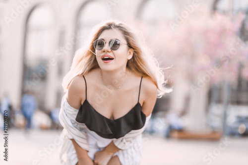 Fototapeta Naklejka Na Ścianę i Meble -  Portrait Beautiful young blonde girl in sunglasses with puffy lips and sexy feminine body posing on  the street  with pink flowers on the background  on sunny day 