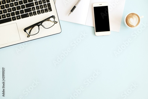Minimalistic flat lay composition of black & white laptop computer keyboard, cell phone gadget, cup of coffee & folded glasses on blue surface desk table background. Workspace top view, copy space