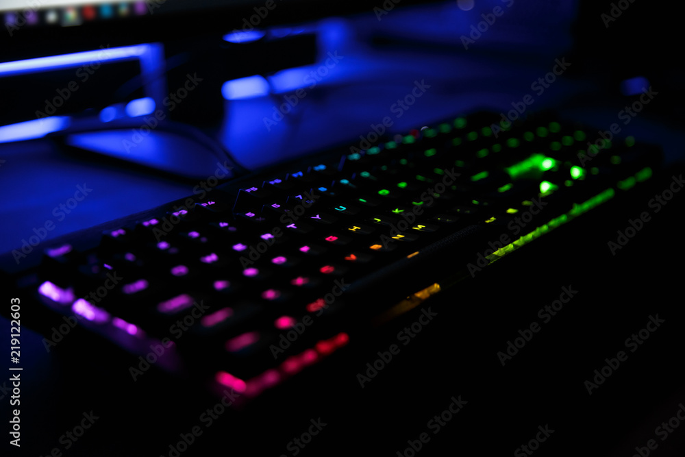 Close up view of workplace with led rainbow backlight gaming keyboard of computer, lying on table in dark room