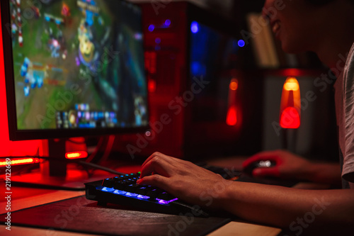 Photo of young gamer guy looking at screen and playing video games on computer in dark room, wearing headphones and using backlit rainbow keyboard