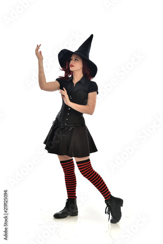 full length portrait of red haired girl wearing black witch costume and pointy hat. standing pose, isolated on white studio background.