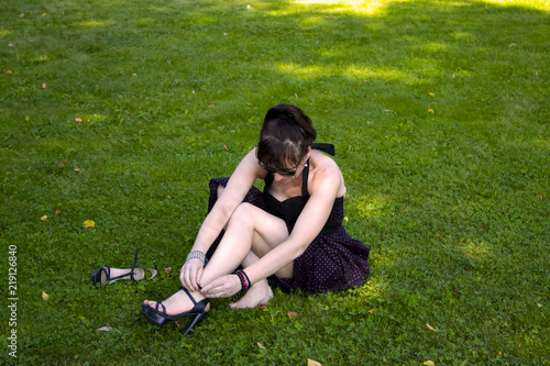 Portrait of a seductive young brunette in high heels smiling, sitting, lying on the green fresh grass in a sunny spring park, in a stylish black dress