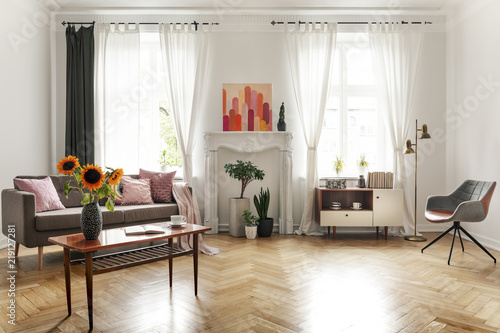 Sunflowers on wooden table next to sofa in spacious flat interior with armchair and poster. Real photo