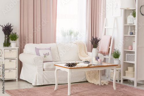 Real photo of bright provencal sitting room interior with white sofa, wooden coffee table on dirty pink carpet, rack with decor and fresh lavender