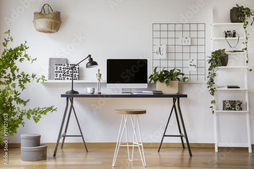 Hairpin stool standing by the wooden desk with mockup computer screen, metal lamp and coffee cup in real photo of white home office interior photo