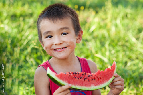Happy asian child eating watermelon in the garden. Kids eat fruit outdoors. Healthy snack for children. Little boy playing in the garden biting a slice of watermelon.