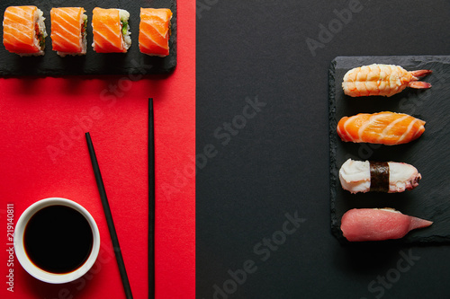 flat lay with soya sauce in bowl, chopsticks and sushi sets on black slate plates on red and black background photo