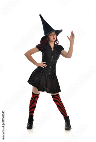 full length portrait of red haired girl wearing black witch costume and pointy hat. standing pose, isolated on white studio background.