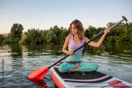 Young woman sitting on paddle board, practicing yoga pose. Doing yoga exercise on sup board, active summer rest