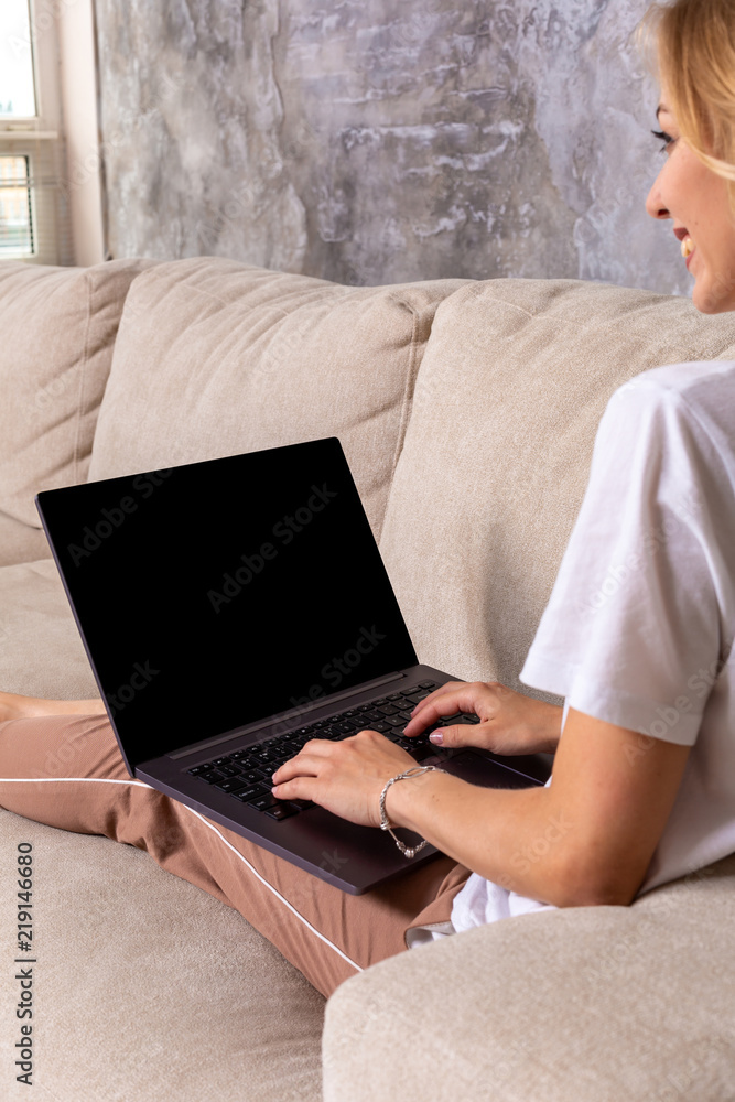 Fototapeta premium Woman on sofa working on laptop with mockup blank screen. Empty copy space on monitor for advertisement. Black screen.