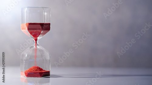 Red sand running through the shape of modern hourglass on white table.Time passing and running out of time. Urgency countdown timer for business deadline concept with copy space