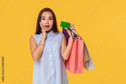 Charming pretty attractive sexy girl showing credit card and holding many colorful packages in hand, it is comfortable to use bank card for shopping, standing over yellow background