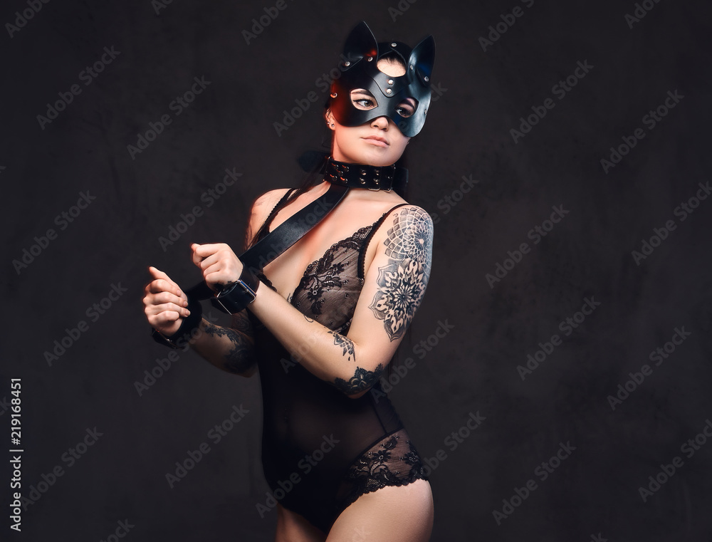 Sexy woman wearing black lingerie in BDSM cat leather mask and accessories posing on dark background. 